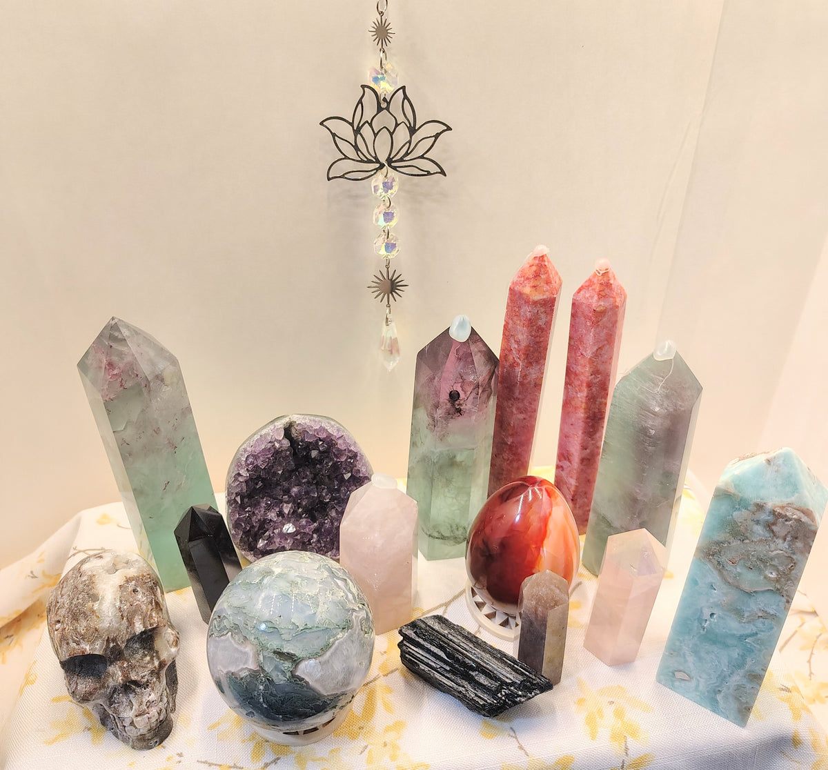 A collection of assorted crystals, quartz points, a geode, a polished blue stone orb, a black obsidian slice, and a skull-shaped crystal arranged on a white fabric-covered surface against a plain light background.